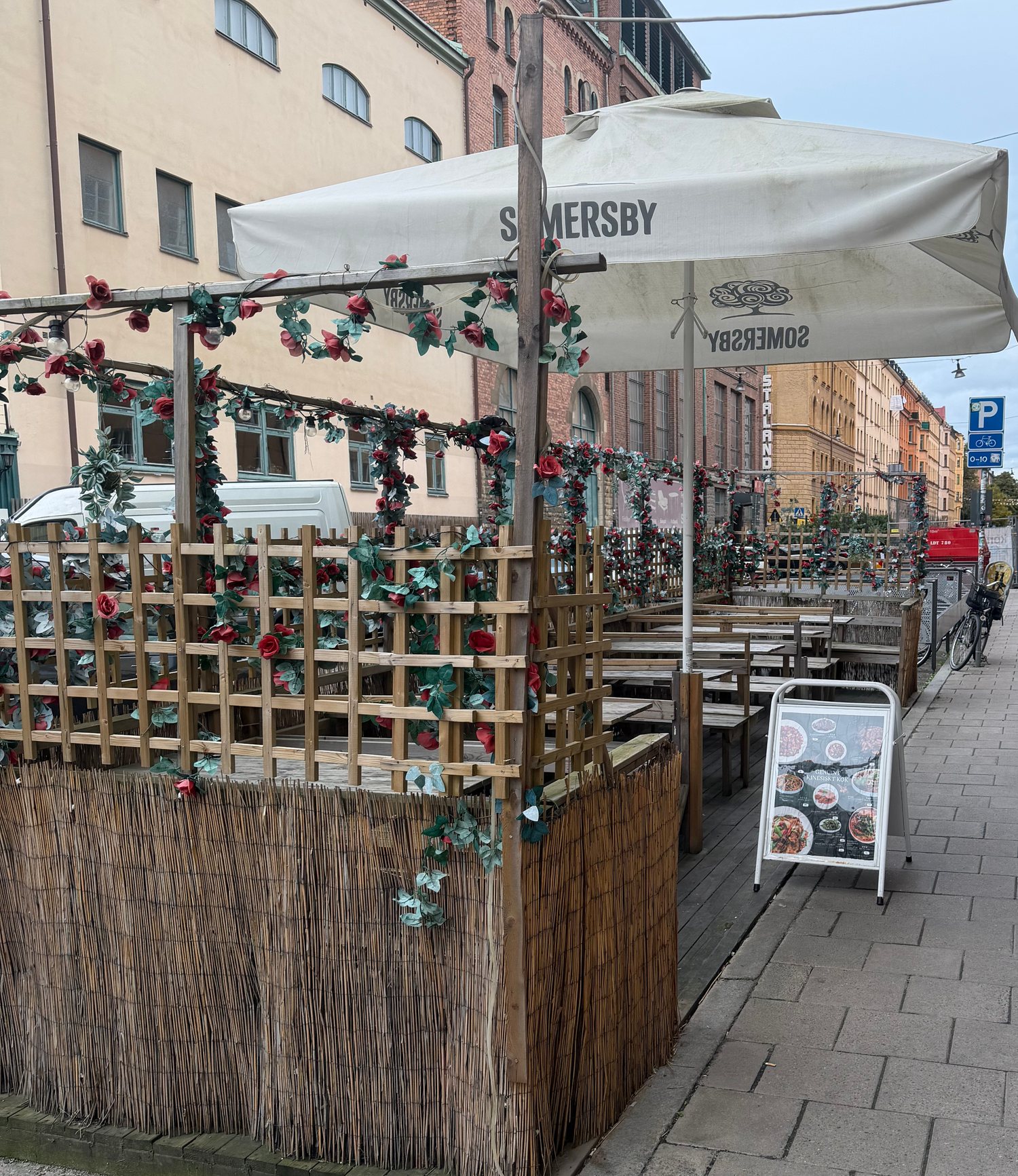 Uteserveringen på Åsögatan med spaljé, rosor, vassstaket och parasoll, och en menyskylt på trottoaren